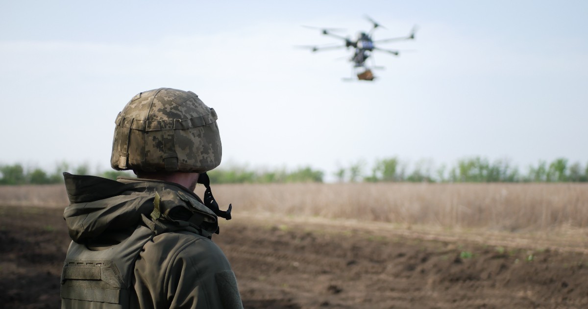 Imaginea surprinde un militar echipat cu cască și vestă de protecție, observând un câmp deschis, în timp ce o dronă zboară la joasă altitudine în fundal.