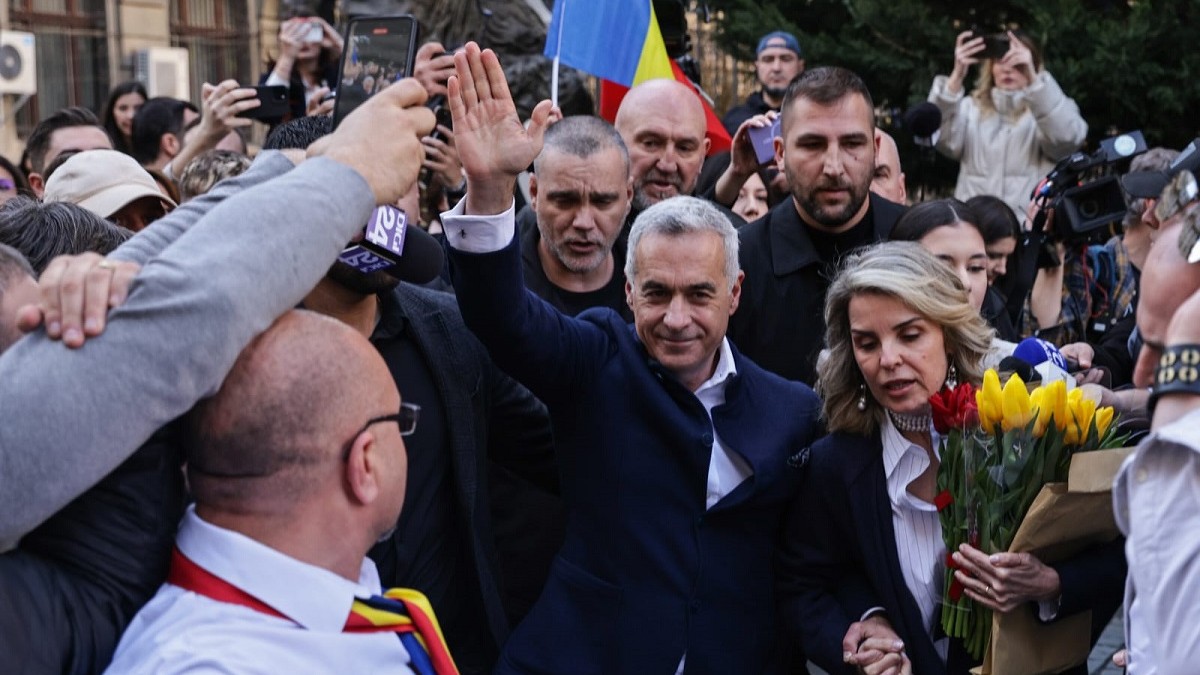 Un bărbat cu mâna dreaptă ridicată în mijlocul unei mulțimi, cu o femeie alături de el, amândoi îmbrăcați în negru. Femeia are un buchet de flori galbene