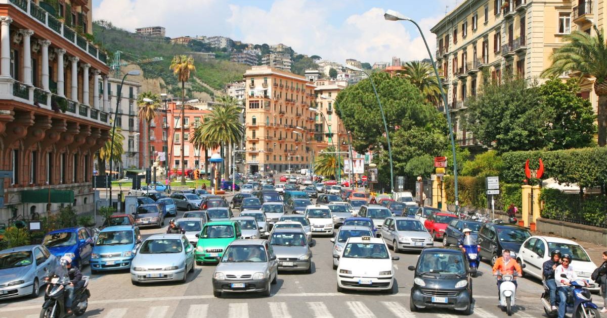 Trafic urban intens într-un oraș din Italia, într-o zonă centrală cu clădiri istorice, palmieri și bulevarde aglomerate. Zeci de mașini – în special modele mai vechi, inclusiv diesel – ocupă toate benzile unei intersecții mari