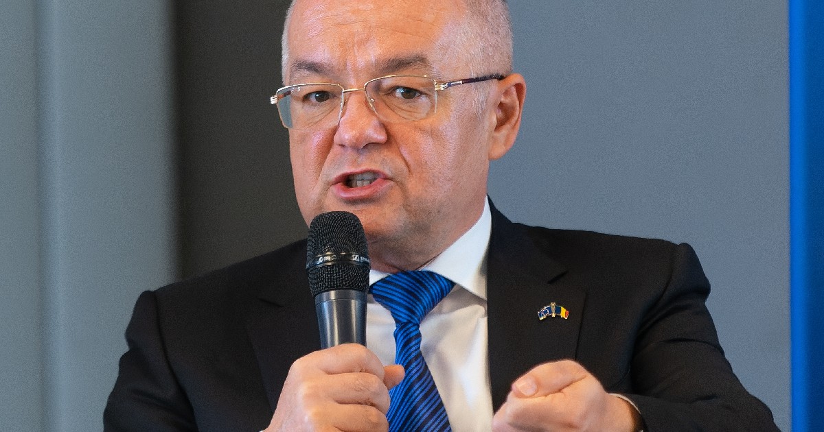 Imaginea prezintă un prim-plan al politicianului Emil Boc, purtând ochelari și costum, în timp ce vorbește la un microfon și gesticulează.