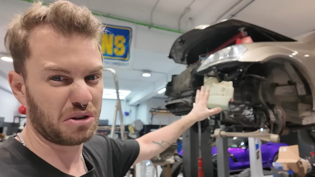 Un bărbat cu o expresie de dezgust, arătând spre zona frontală dezasamblată a unei mașini ridicate pe o platformă într-un atelier auto