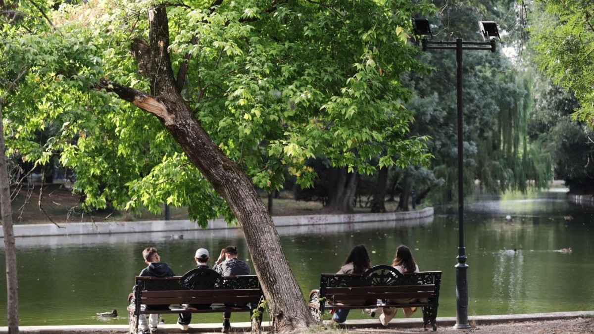 Două bănci, una lângă alta, pe care stau băieți și fete, cu spatele, iar în fașța lor se vede lacul Cișmigiu și pomi înfrunziți, parcul Cișmigiu din București, 30 septembrie 2025