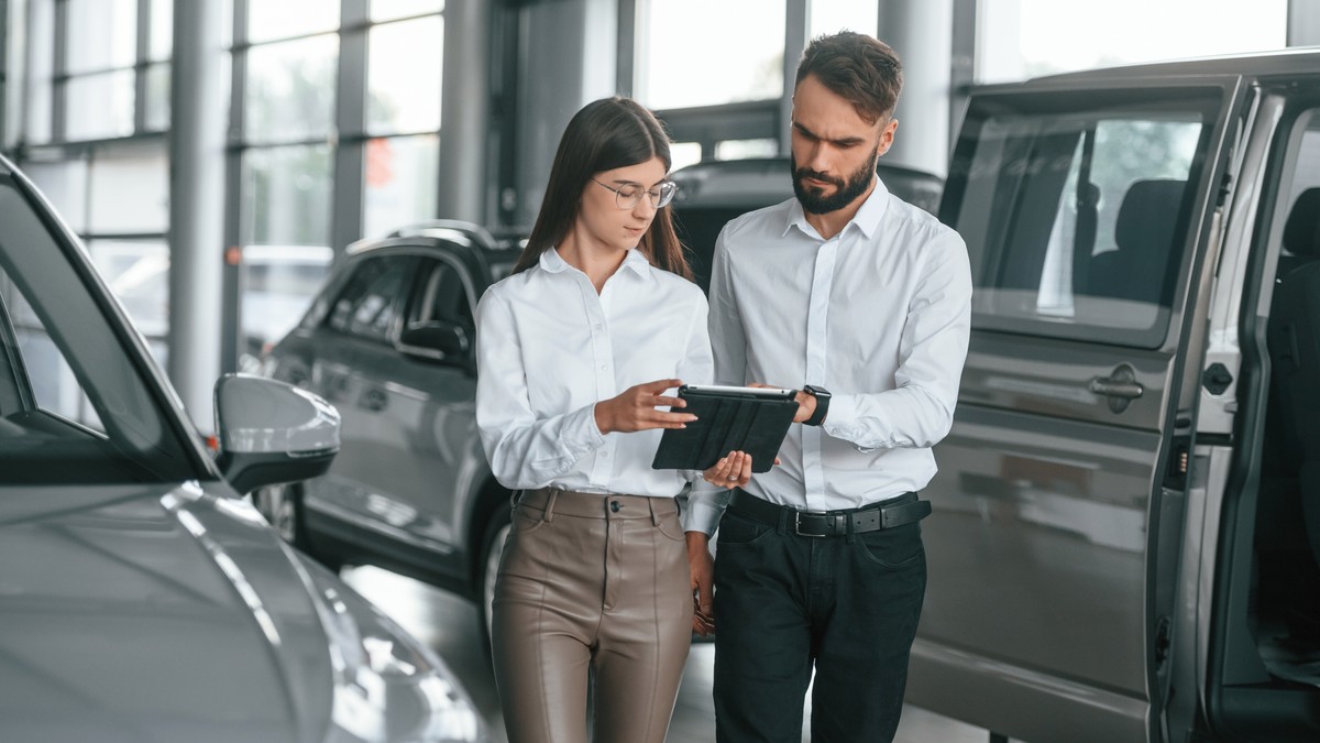 O femeie si un barbat, imbracati business, intr-o reprezentatnta auto. In jurul lor sunt masini gri