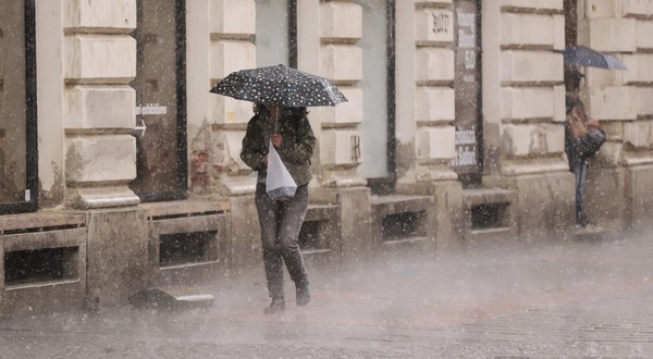 O persoană adăpostindu-se sub o umbrelă cu buline în timp ce merge pe trotuar pe o ploaie torențială, lângă fațada unei clădiri istorice.