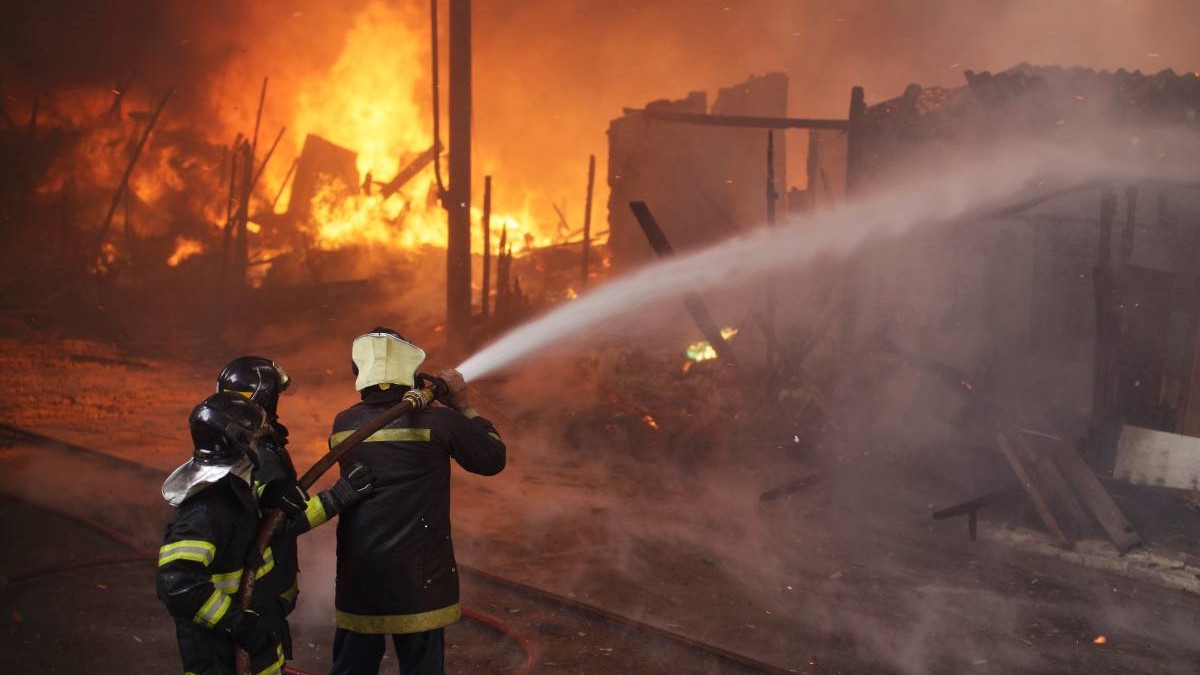 Imagine de noapte, trei pompieri cu un furtun cu apa incearca sa stinga un incendiu care arde cu flacara mare si fum