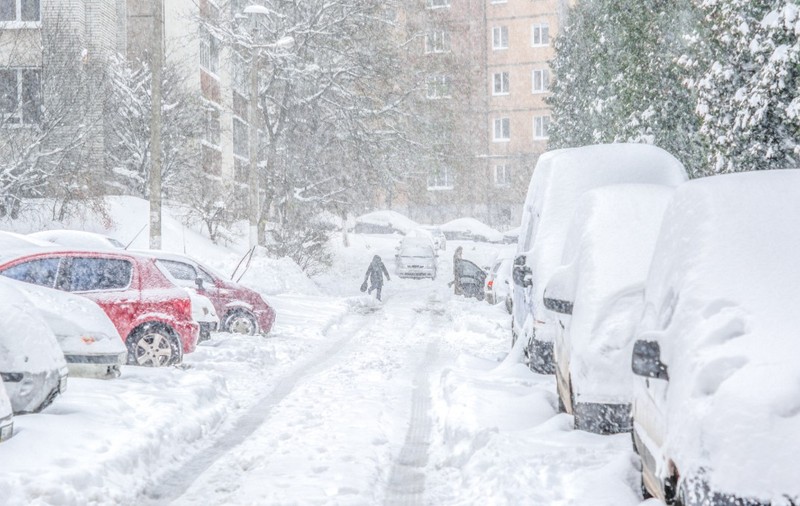 O stradă dintre blocuri acoperită de un strat gros de zăpadă în timpul unei ninsori abundente, cu numeroase autoturisme parcate pe margine și o persoană care merge pe aleea îngustă curățată.