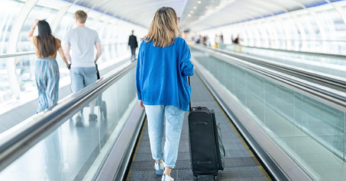 Femeie îmbrăcată în pulover albastru, blugi si adidasi, mergând cu valiza pe banda rulantă într-un aeroport