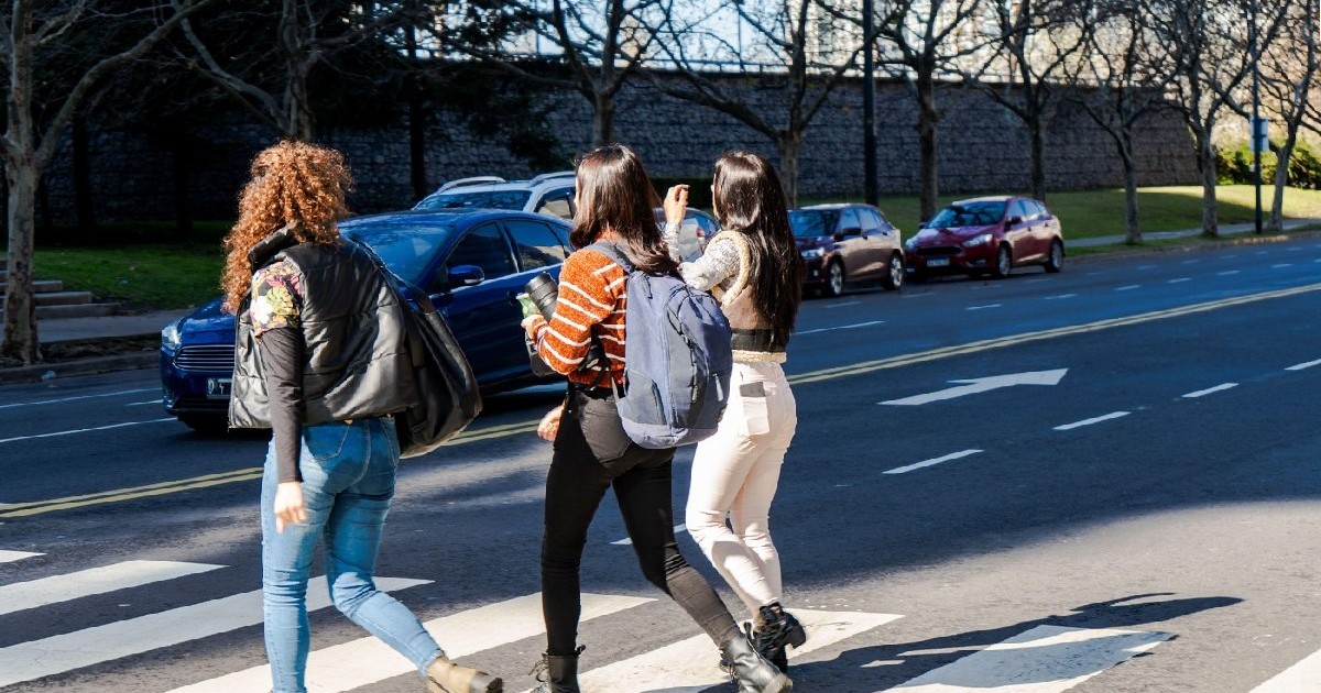 Trei tinere studente traversează o trecere de pietoni în oraș într-o zi însorită.