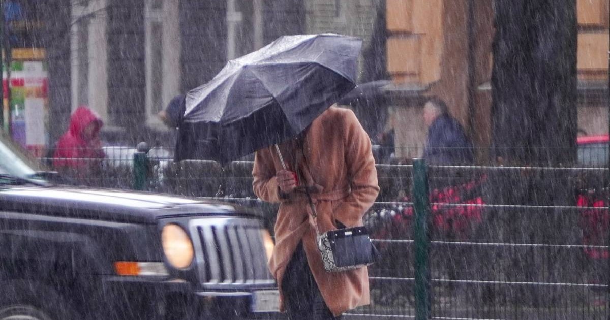 Imaginea surprinde o persoană văzută din fata, cu un palton crem, care se protejează de vant si ploaie sub o umbrelă neagra, într-un cadru urban, se vede o masina neagra cu farurile aprinse, mai multe persoane cu pelerine de ploaie in spate, ploua torential
