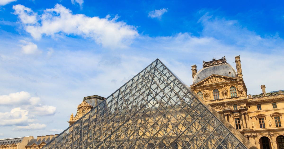 Imaginea prezintă celebra Piramidă de Sticlă din fața Muzeului Luvru din Paris, care contrastează puternic cu arhitectura clasică, masivă, a clădirilor din jur, sub un cer albastru cu nori puțini. Piramida modernă, cu structura sa metalică și panourile de sticlă, domină centrul cadrului, în timp ce la bază se pot observa câțiva vizitatori.