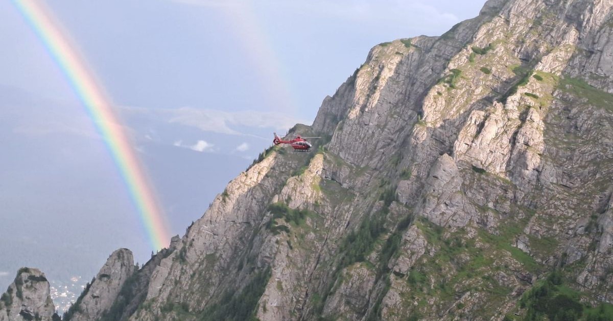 un peisaj montan abrupt și impunător. Partea dreaptă a cadrului este dominată de un versant stâncos masiv, cu pante foarte înclinate, brăzdat de crăpături adânci și presărat pe alocuri cu petice de vegetație montană. Chiar în apropierea crestei ascuțite a acestui munte, un elicopter este surprins zburând la joasă înălțime, silueta sa părând mică în comparație cu relieful accidentat din fundal. În partea stângă a imaginii se deschide o prăpastie adâncă, mărginită la bază de câteva vârfuri stâncoase mai ascuțite, peste care se arcuiește vizibil curba amplă a unui curcubeu. În depărtare, dincolo de hău, se întrezăresc contururile altor lanțuri muntoase.