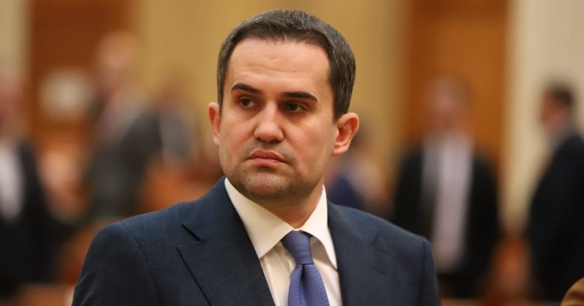 Ciprian Constantin Șerban, cu o grimasă serioasă și încrutat, purtând o cămașă albă, un sacou bleu-marin și o cravată albastru deschis