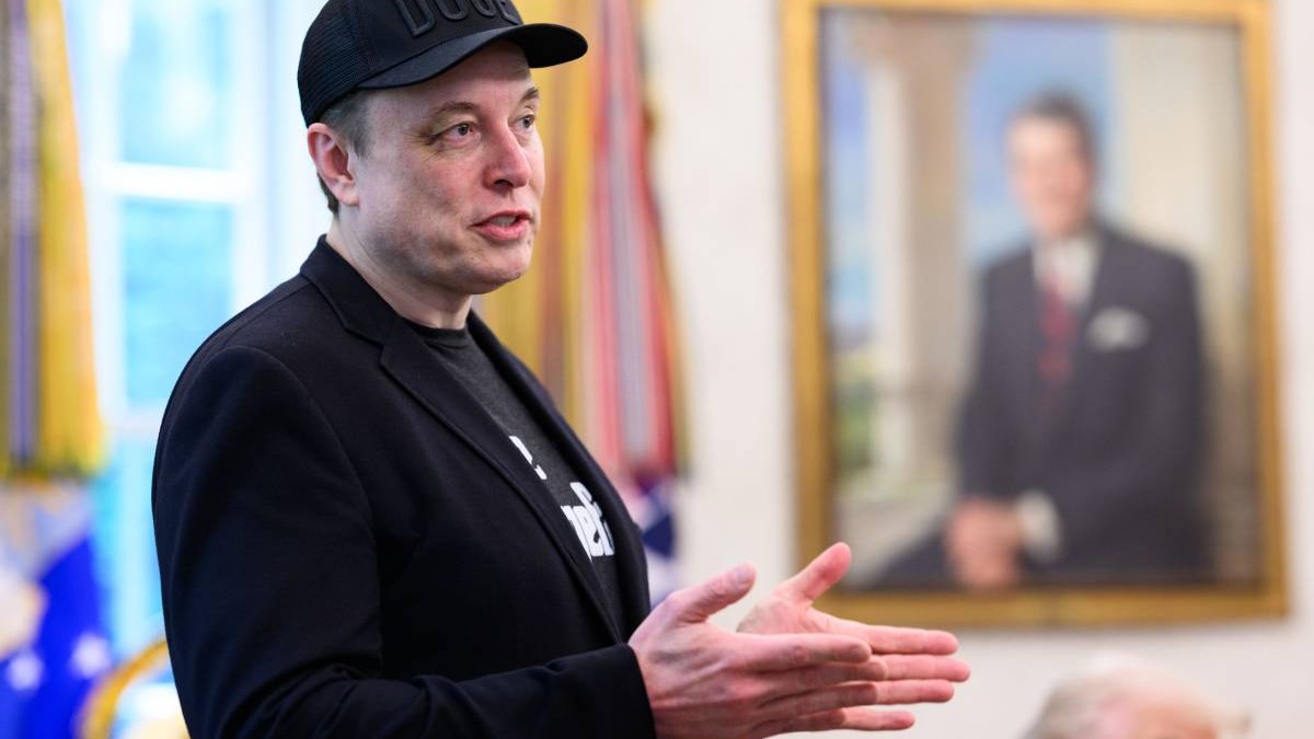 Elon Musk în Biroul Oval de la Casa Albă, poartă o șapcă neagră pe cap, gesticulează cu palmele ridicate și are un ochi vânăt