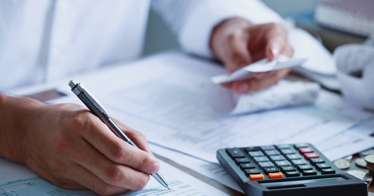 Un bărbat care stă la birou, îmbrăcat cu o cămașă albă, în timp ce calculează taxele și impozitele și notează totul pe foi