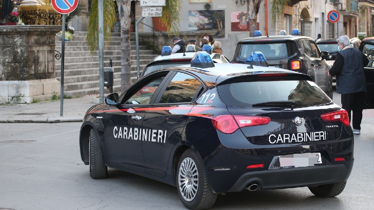 În imagine este o mașină neagră de poliție pe scrie CARABINIERI, staționată pe o stradă aglomerată, în fața unei clădiri, cu un semafor și indicatoare rutiere în fundal.