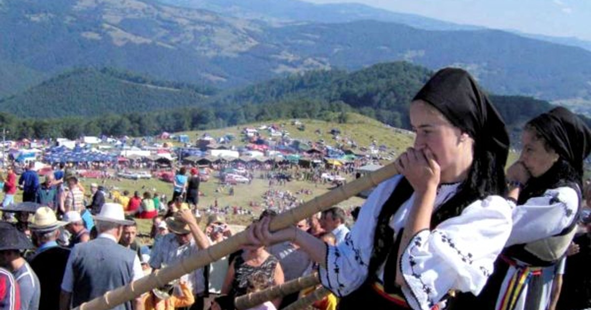 Târgul de Fete de pe Muntele Găina 2018, LIVE pe internet. Fotografie din 2017, de la Târgul de fete de pe Muntele Găina, cu mai multe tinere care suflă în bucium și mulți spectatori pe lângă ele