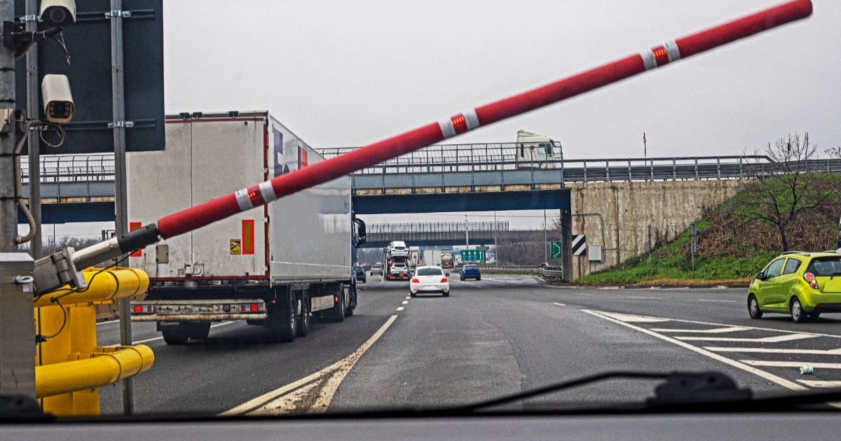 În imagine este prezentată perspectiva din interiorul unui vehicul aflat la o stație de taxare, surprinzând o barieră roșie cu dungi albe care se ridică pentru a permite accesul pe o autostradă.