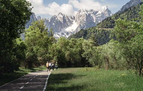 Cicliștii pedalează de-a lungul traseului ciclist Alpe-Adria, în apropiere de Tarvisio, înconjurați de vegetație luxuriantă și munți înzăpeziți Traseul pitoresc oferă o experiență în aer liber li