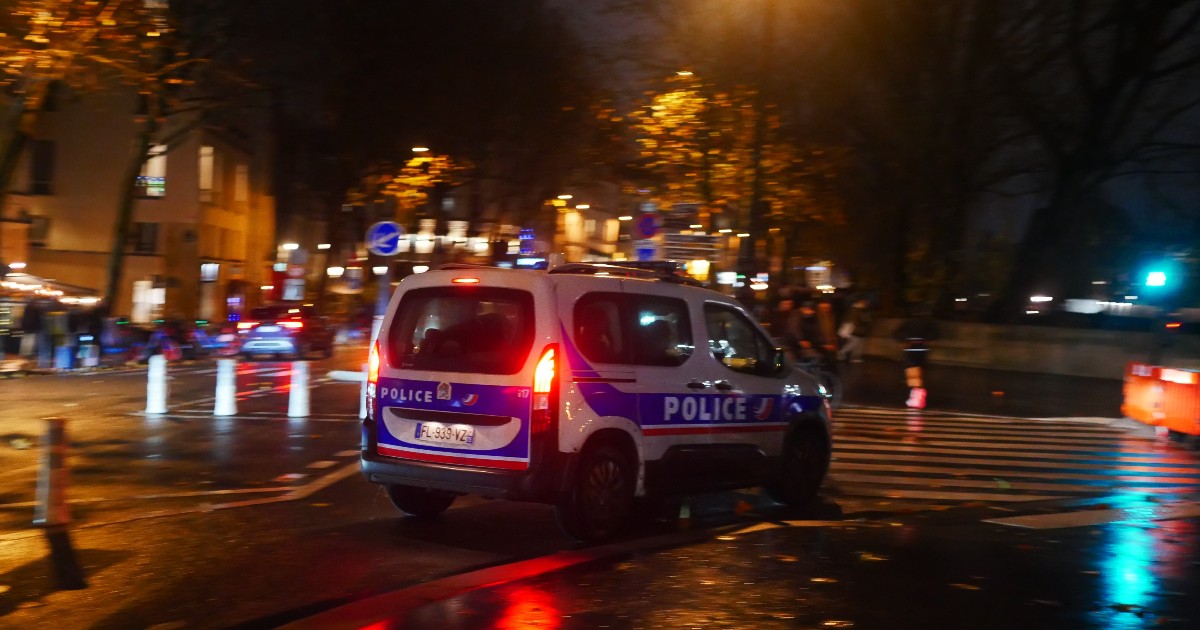 Imaginea surprinde o mașină a poliției franceze care circulă noaptea pe o stradă urbană, cu luminile aprinse, pe carosabil umed, reflectând culorile semafoarelor și ale iluminatului stradal. Atmosfera este una tensionată și sobră, sugerând o intervenție sau o anchetă aflată în desfășurare.