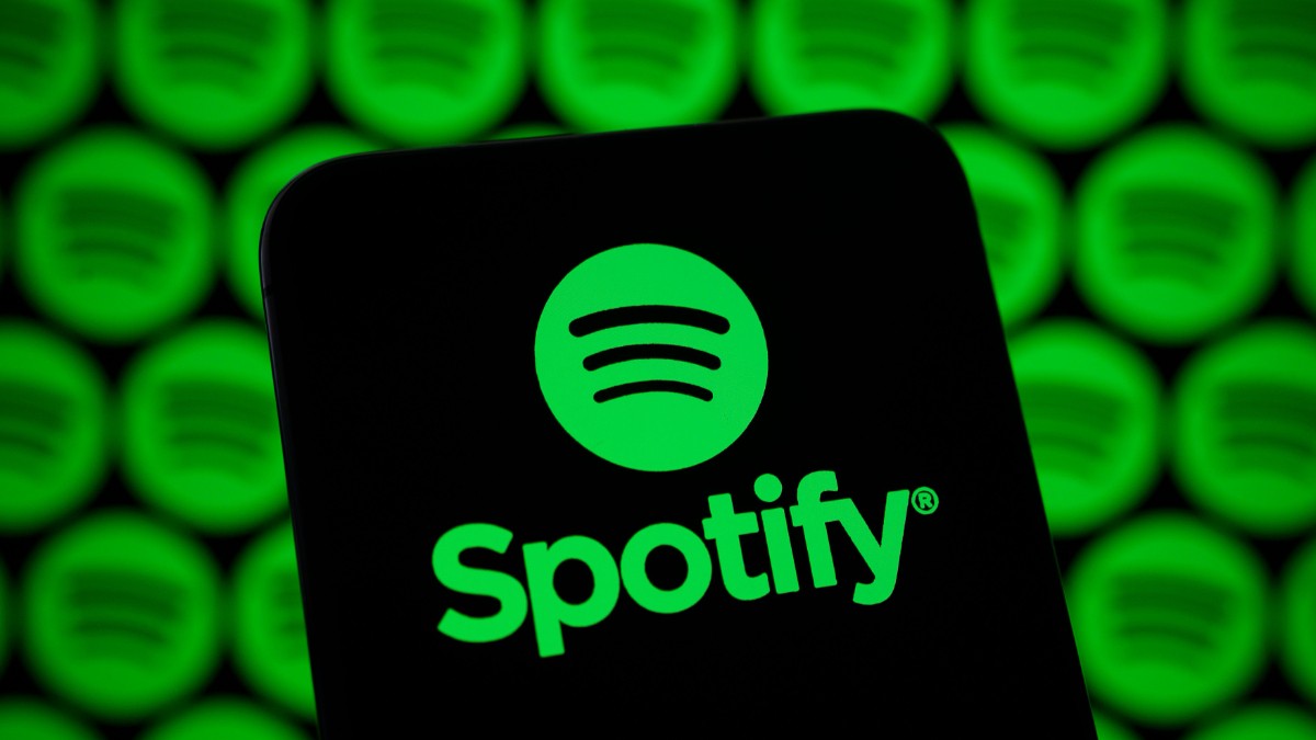 În imagine apare logo-ul Spotify afișat pe ecranul unui dispozitiv mobil, în prim-plan. Fundalul este dominat de mai multe logo-uri Spotify verzi, repetate și estompate.