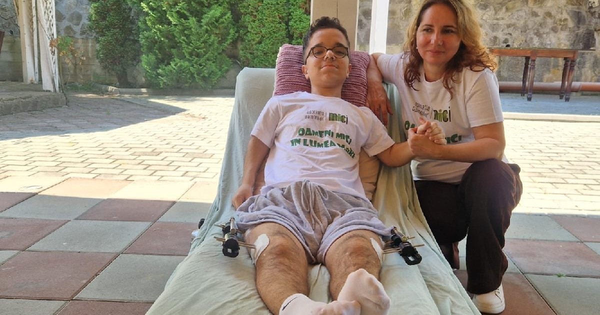 Un baiat sta intains pe un pat pliant, este pansat la picioare si are fixatoare in picioare. Langa el e mama sa. A venit in tabara dupa o operatie de acondroplazie