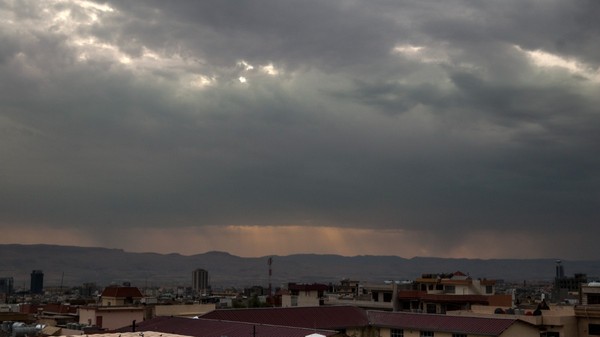 Vedere panoramică asupra unui oraș cu clădiri și dealuri în fundal, sub un cer acoperit de nori groși și întunecați de furtună, prin care pătrund câteva raze de lumină la orizont.