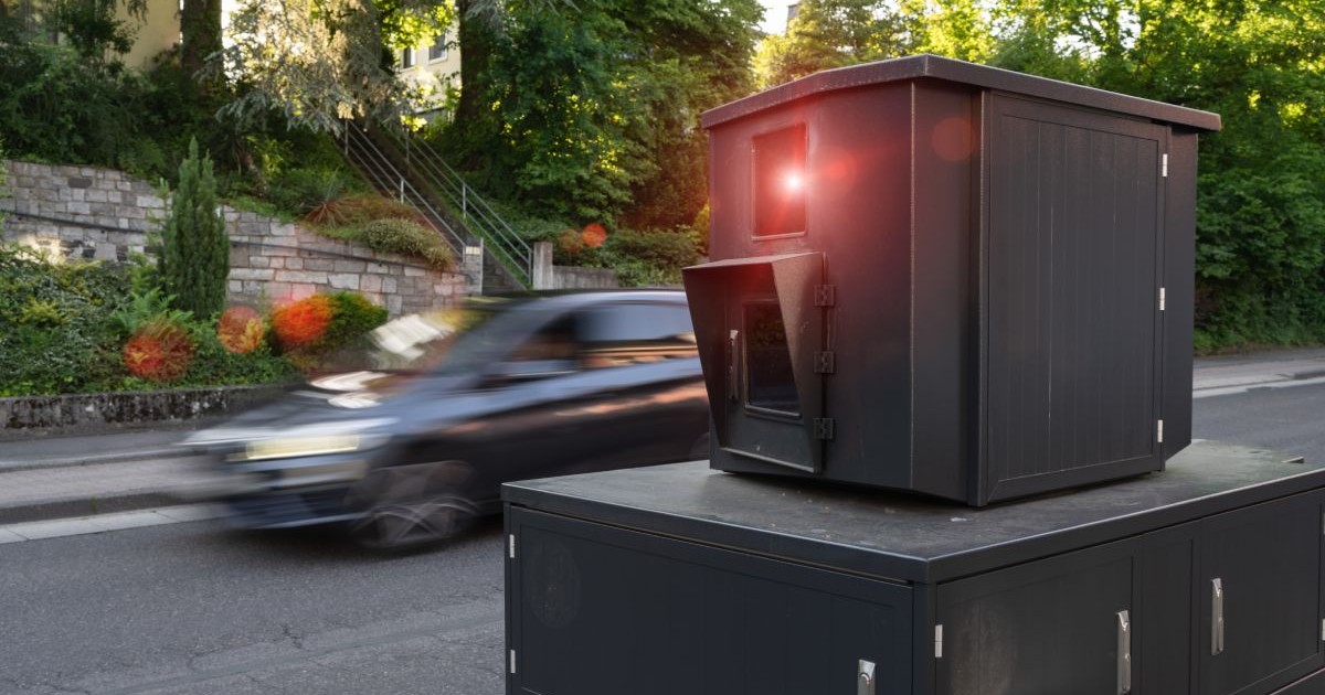 Un radar mobil al Poliției, de culoare neagră, cu o cameră și un senzor infraroșu, lângă o șosea, și o mașină de culoare închisă care circulă