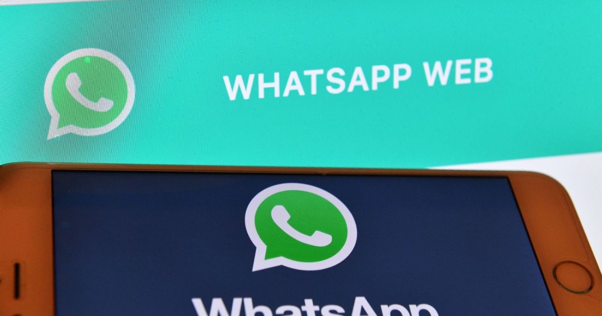 Logo-ul Whastapp afișat pe ecranul unui telefon, care este ținut în fața unui fundal de browser Google Chrome cu site-ul WhatsApp Web deschis.