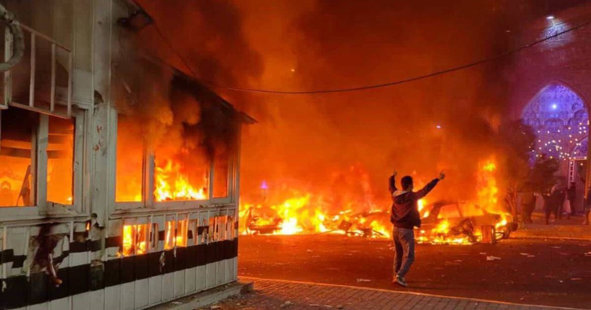 Imagine de noapte din timpul protestelor din Iran, se vad flacari mari in intuneric, care au cuprins totul, silueta unei persoane cu mainile ridicate in mijlocul lor