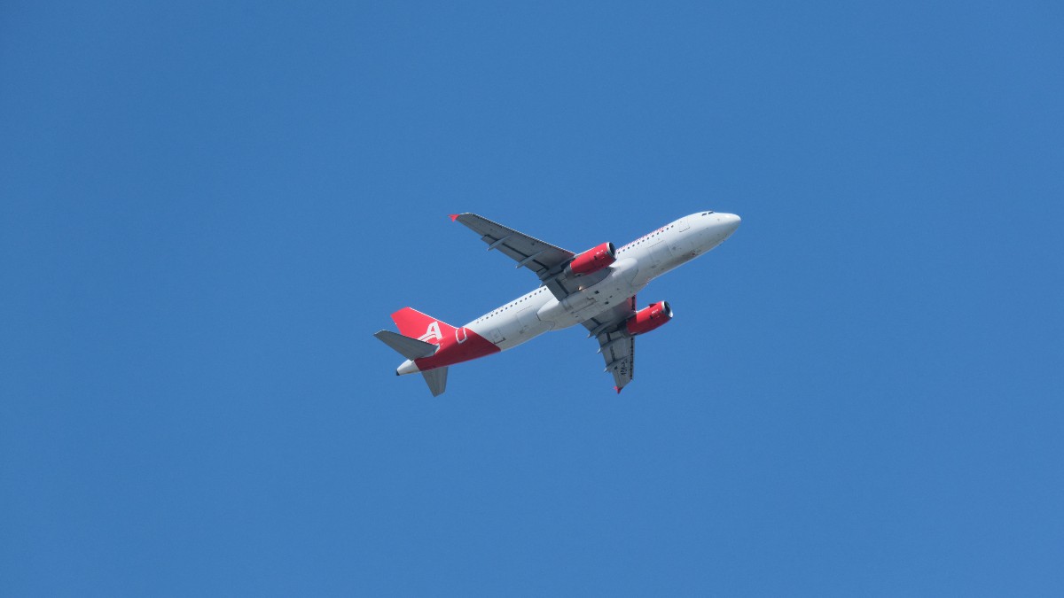 Imaginea surprinde un avion de pasageri bimotor de culoare albă, cu accente roșii pe aripile motorului și pe stabilizatorul vertical, în plin zbor pe un fundal de cer albastru senin. Avionul este fotografiat de jos, cu trenul de aterizare retractat, sugerând o ascensiune sau croazieră la altitudine joasă.