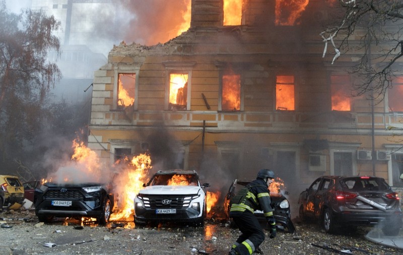 O clădire rezidențială istorică și mai multe autoturisme parcate în fața ei cuprinse de flăcări violente, în timp ce un pompier echipat de intervenție acționează la fața locului.