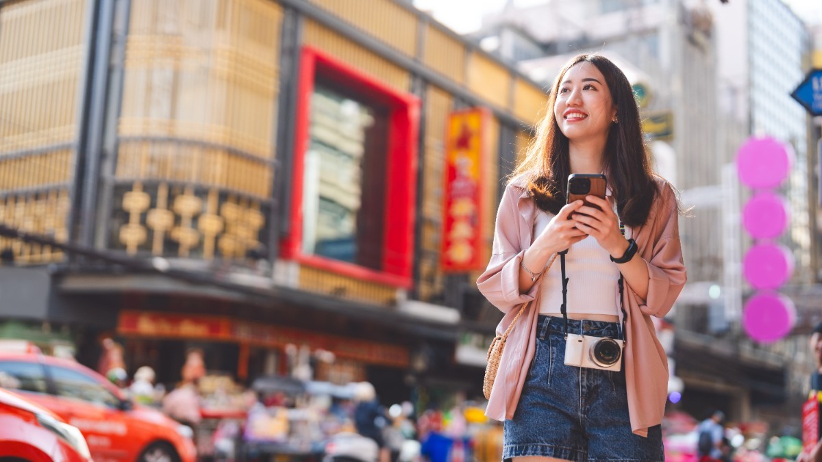 O femeie asiatică tânără cu un telefon în mână, pe o stradă din Hong Kong, în timp ce face o fotografie în fața unei clădiri înalte