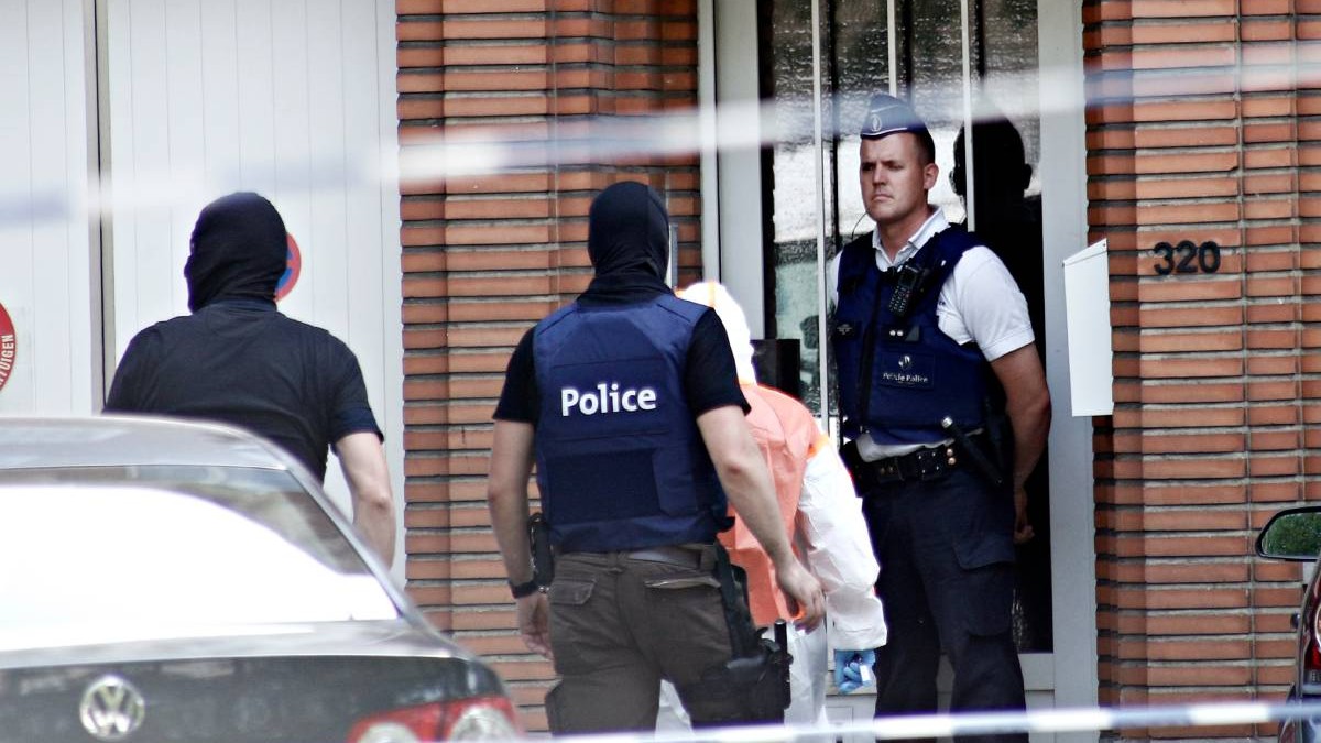 Imagine cu 4 persoane, 3 in uniforma de politi, o a patra cu un combinezon alb si galben, in fata unei case, Belgia