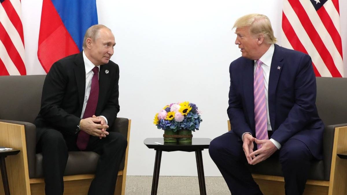 vladimir putin si donald trump in timpul unui dialog. ambii sunt asezati pe scaun si intre ei este o masa rotunda. in spatele lor se afla stagul rusiei si al statelor unite ale americii