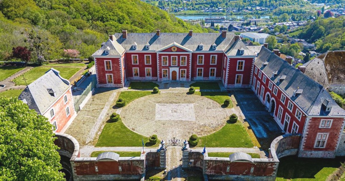 Imaginea prezintă o vedere aeriană a unui castel cu fațadă roșie și detalii albe, dispus în formă de U în jurul unei curți interioare amenajate cu gazon circular, situat într-un cadru natural deluros.