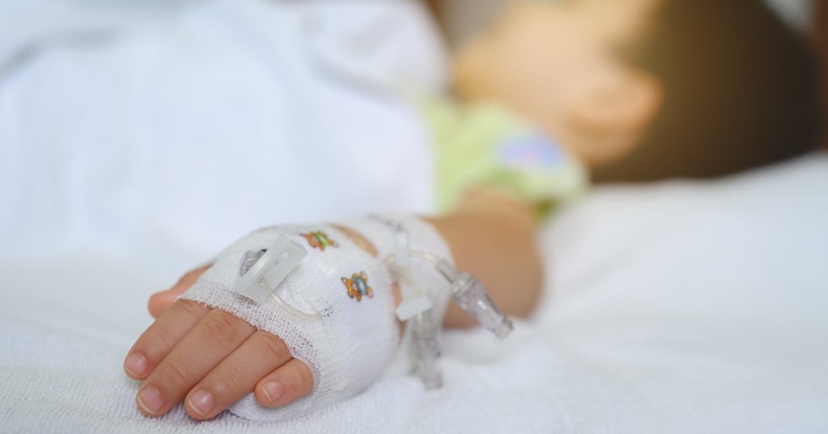 Imaginea surprinde mâna unui copil aflat pe un pat de spital, cu o branulă fixată și bandajată, semn al unei intervenții medicale. Fundalul este estompat, accentuând starea de vulnerabilitate și contextul clinic.