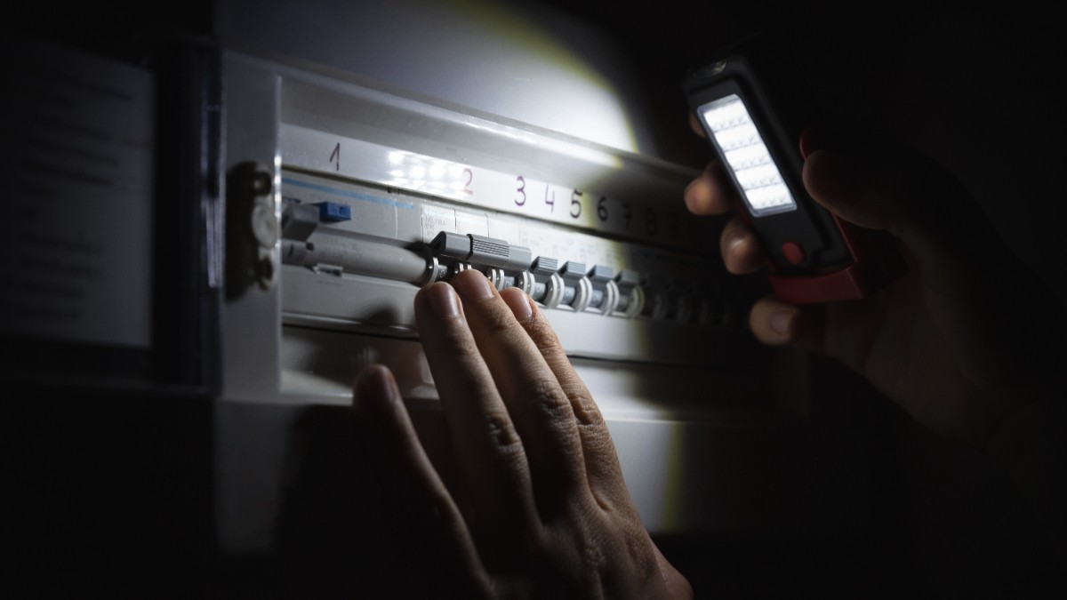 Un bărbat verifică cu lumina de la telefonul mobil un panou electric într-un mediu întunecat și iluminează siguranțele și întrerupătoarele din panou.