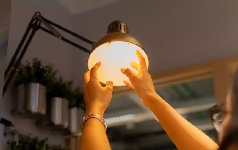 O femeie înșurubează un bec cu mâinile într-o veioză și se vede lumina. Foto: Shutterstock