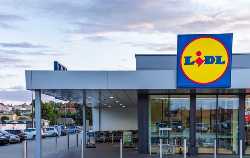 Un magazin Lidl modern este surprins din exterior, cu intrarea largă și logo-ul vizibil deasupra ușilor automate. În fața clădirii se află parcarea și zona pentru cărucioare, sugerând acces facil pentru clienți.
