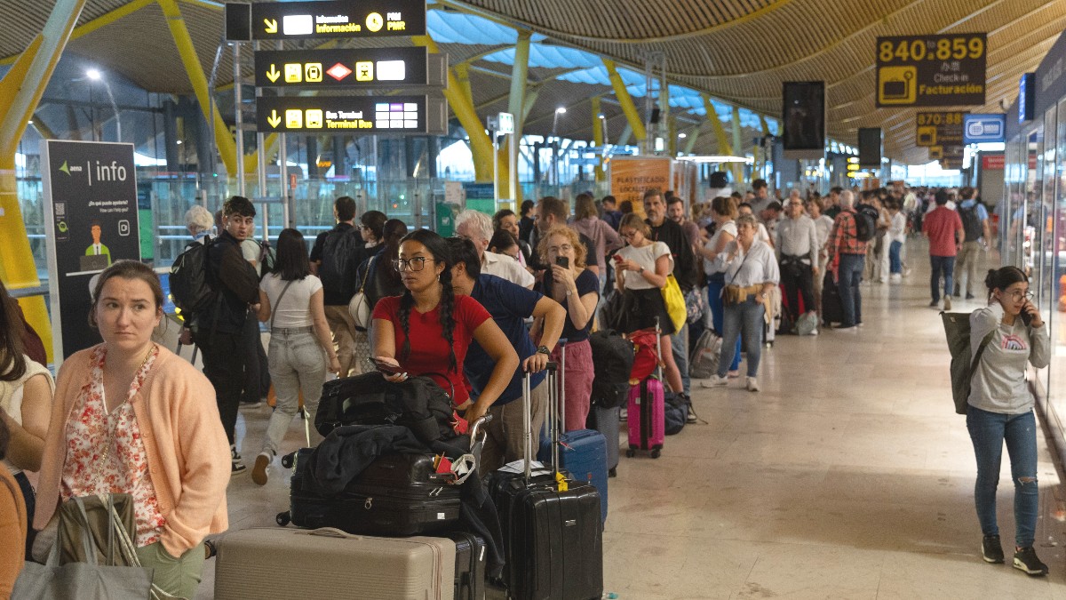 Imaginea înfățișează numeroși pasageri cu bagaje care așteaptă la coadă în terminalul aglomerat al unui aeroport din Spania.