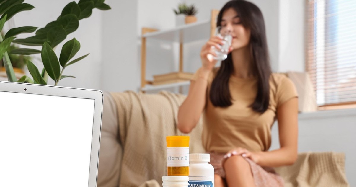 Tânără luând pastile acasă, pe masă se văd recipiente cu diferite vitamine B și laptopul