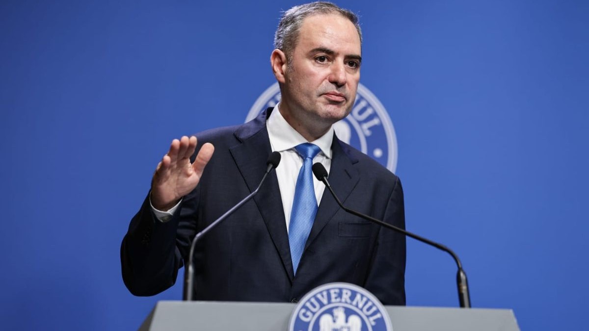 Alexandru Nazare, îmbrăcat într-un sacou negru, o cămașă albă și o cravată albastră, în timpul unei conferințe de presă de la Palatul Victoria, 13 august 2025