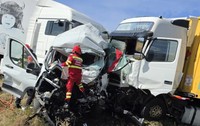 Imaginea prezintă urmările unui accident grav, în care o dubă albă este zdrobită de două TIR-uri, în timp ce un echipaj de pompieri și paramedici SMURD intervine la fața locului.