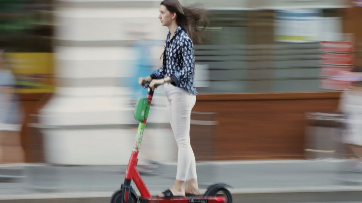 O tânără îmbrăcată în pantaloni albi, o cămașă bleumarin cu alb și șlapi negri merge pe o trotinetă electrică, pe o stradă din București, august 2025