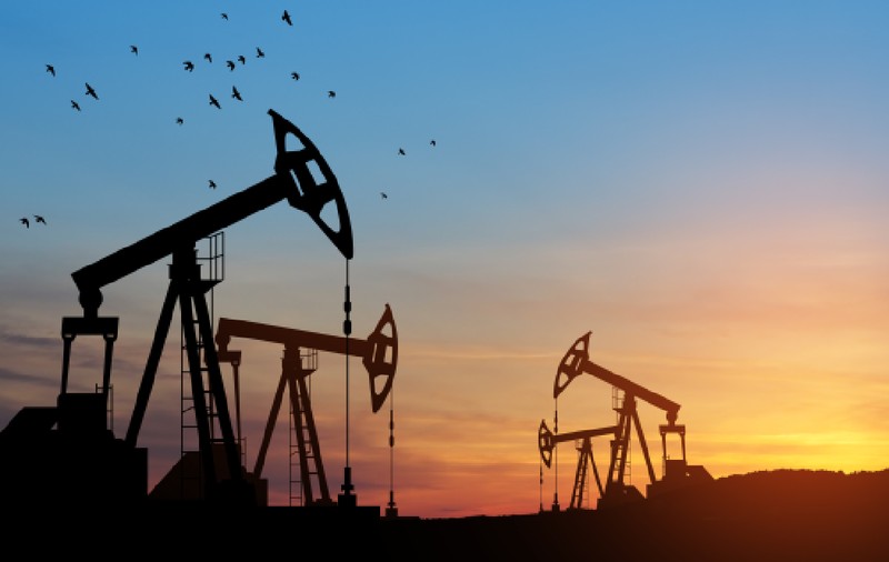Imaginea prezintă mai multe sonde de petrol aflate în funcțiune pe fundalul unui apus de soare colorat, în timp ce un stol de păsări zboară pe cerul de deasupra lor.