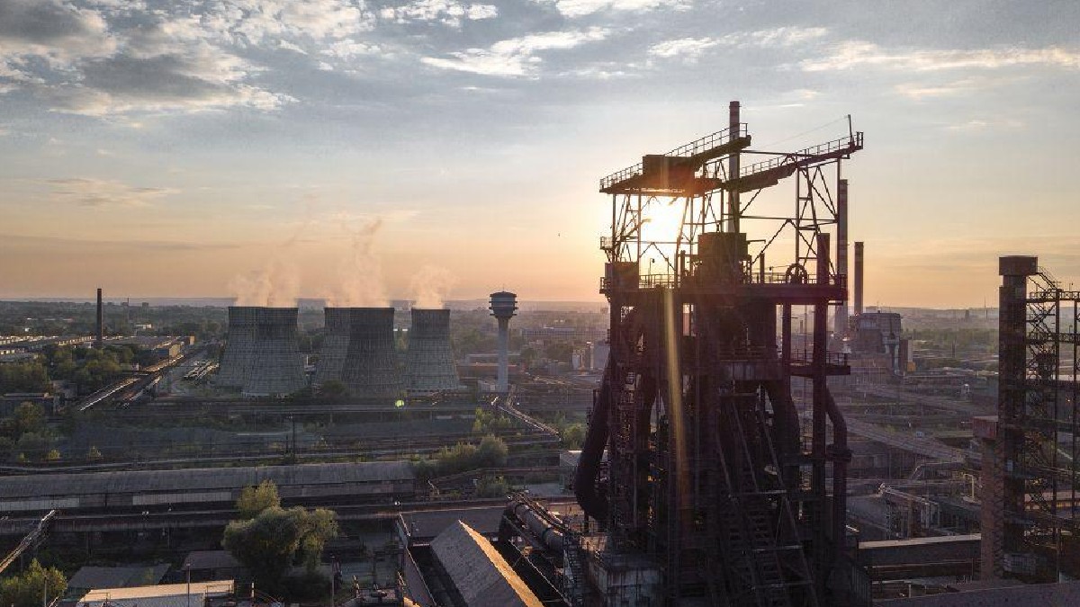 Imaginea aeriană surprinde un peisaj industrial vast, dominat în prim-plan de structura metalică masivă a unui furnal sau a unei cocserii, silueta fiind conturată de lumina puternică a apusului. În planul îndepărtat, se disting turnuri de răcire înalte, din care iese abur, și un turn de apă, confirmând că scena reprezintă Combinatul Siderurgic Liberty Galați.