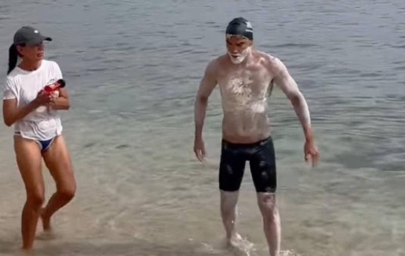 Spyros Chrysikopoulos iese din mare la capătul traversării dintre Grecia și Italia, după aproape 32 de ore de înot. Sportivul poartă cască și ochelari de înot, iar corpul îi este acoperit de cremă de protecție. Lângă el se află o femeie, pe mal, în timp ce acesta pășește prin apa mică spre plajă.