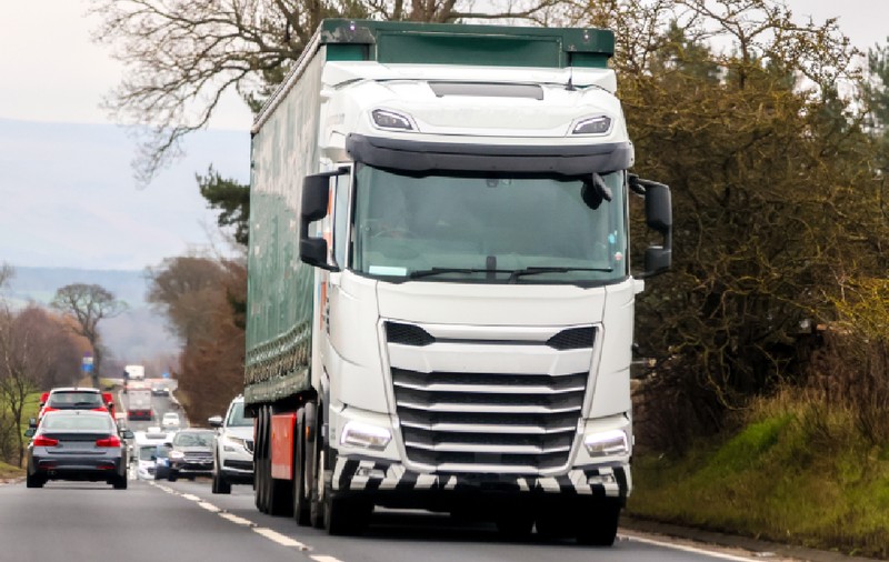 O imagine dinamică ce surprinde un camion alb cu remorcă verde circulând pe o șosea cu două benzi, fiind urmat de o coloană de autoturisme într-un peisaj rural.