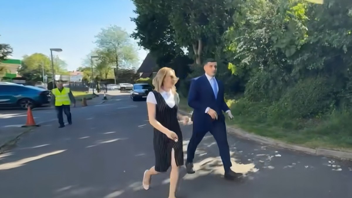 George Simion, îmbrăcat în costum bleumarin, cămașă albă și cravcată bleu, alături de o femeie îmbrăcată într-un sarafan negru și cu tricou alb, mergînd pe o alee