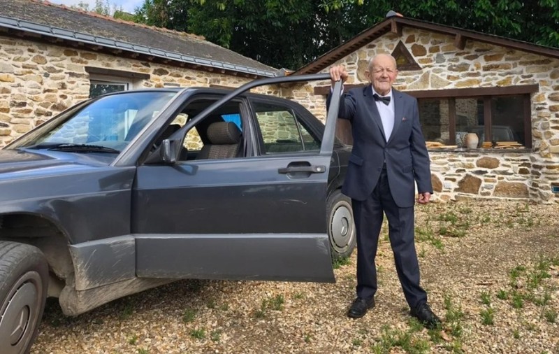 Un bărbat în vârstă, îmbrăcat elegant în costum cu papion, stând lângă portiera deschisă a unei mașini, în fața unei case din piatră.